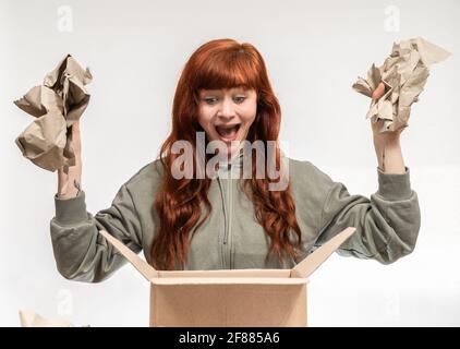 Portrait of a woman with red hair infront of white background with wrapping paper in hands while unboxing a package and having an excited face Stock Photo