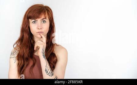 Portrait of tattooed woman with red hair making thinking gesture infront of white background with space on the right Stock Photo