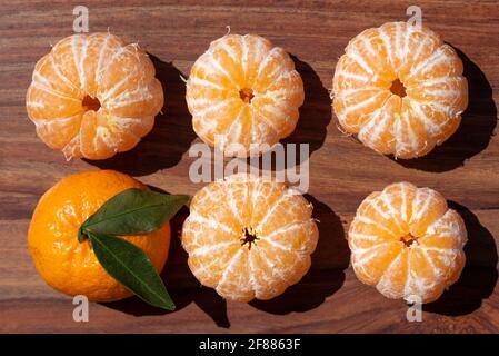 Several tangerines without peel and one tangerine with peel and green leaves lie next to each other on wood and are photographed from above Stock Photo