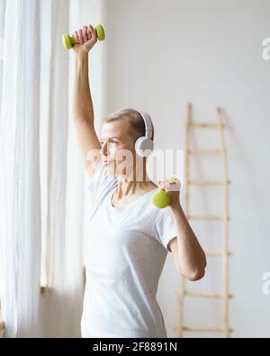 Portrait of smiling senior woman exercising fitness with dumbbells at home. Mature female build up biceps, train arms triceps. Sport for elderly lady. Well-being, wellness for retired female, vertical Stock Photo