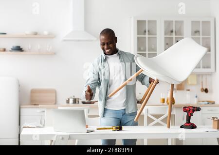 Happy black man showing new chair and thumb up, having video chat on laptop, standing in kitchen, copy space Stock Photo