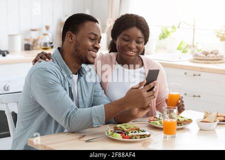 Happy African American Spouses Using Smartphone While Having Breakfast In Kitchen Stock Photo