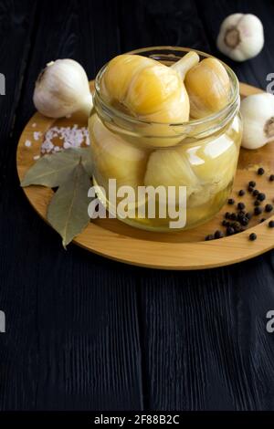 Pickled garlic in the glass jar on the brown cutting board on the black wooden background.Homemade fermented product. Stock Photo