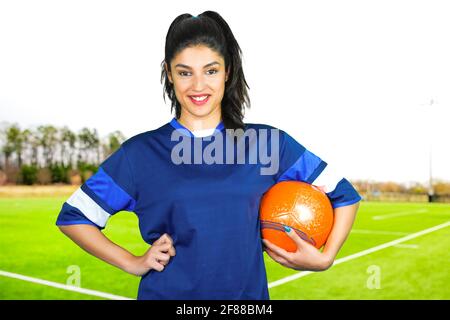 A young soccer player holds her ball under her arm in front of the soccer field. Subject on a blurred background. Perfect shot for emancipation, women Stock Photo