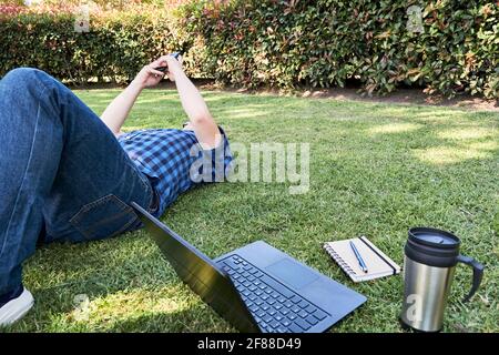 Unrecognizable young man lying on the grass using a cell phone, resting from studying or working outdoors. Next to him, his laptop, a notebook with a Stock Photo