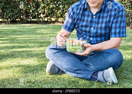 Unrecognizable young man sitting on the grass holding a glass container with a fresh and colorful salad. Healthy living and eating concepts. Stock Photo
