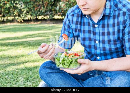 Unrecognizable young man sitting on the grass eating a fresh and colorful salad held in a glass bowl. Healthy living and eating concepts. Close up ima Stock Photo