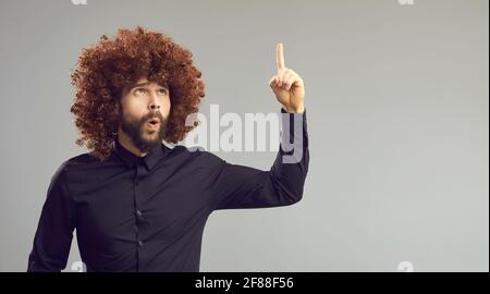 Funny man in big curly wig pointing finger up isolated on gray copy space background Stock Photo