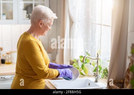 Senior lady washing dishes in kitchen, using rubber gloves Stock Photo