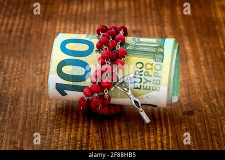 A Catholic cross, a rosary with beads and roll of Euro currency lie on a dark brown wooden table. Relationship of religion to money, faith, spirituali Stock Photo