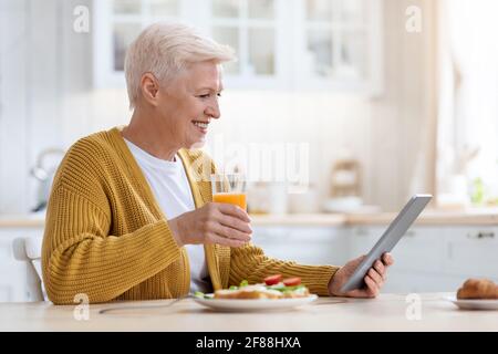 Joyful senior lady with digital tablet having lunch, side view Stock Photo