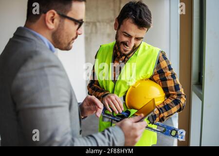 Smiling construction worker standing with supervisor and looking blueprints on tablet while standing in building in construction process. Stock Photo