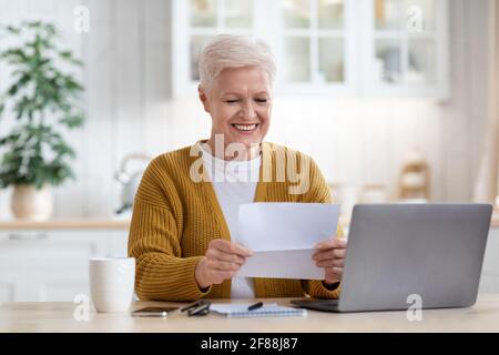 Positive grandmother sitting in kitchen, reading letter Stock Photo