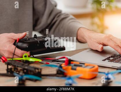 Service man sitting by the table with repairing tools, using laptop and holding quadcopter control panel in his hands Stock Photo