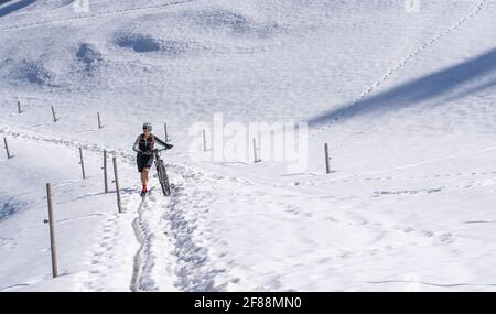 senior woman mountainbiking and pushing her e-mountainbike through a snow spot in early spring, in the Allgaeu Area, Germany Stock Photo