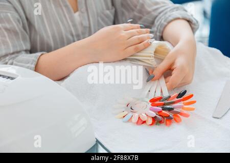 Female hands with artificial acrylic nails picks up new nail polish color during manicure procedure. Colored nail polish process on manicure in beauty Stock Photo