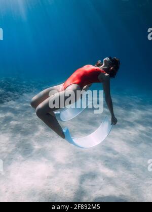 Freediver in red swimsuit with fins posing underwater over sand in tropical ocean. Stock Photo