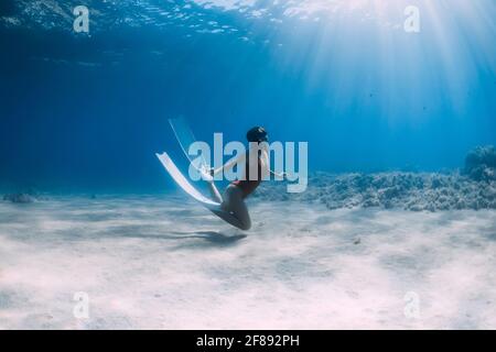 Woman freediver in swimsuit with fins glides underwater over sand in tropical ocean. Stock Photo