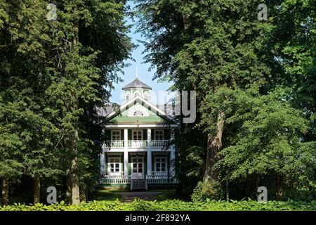 Old green wooden house with a balcony and white columns framed by trees in the manor park. Stock Photo