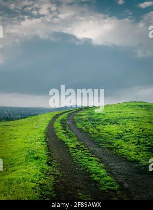 Empty rough road or path in the countryside hill and green grass with cloudy sky during evening in the rainy season. Stock Photo