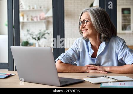 Middle aged woman sitting at home with computer smiling dreamingly. Stock Photo