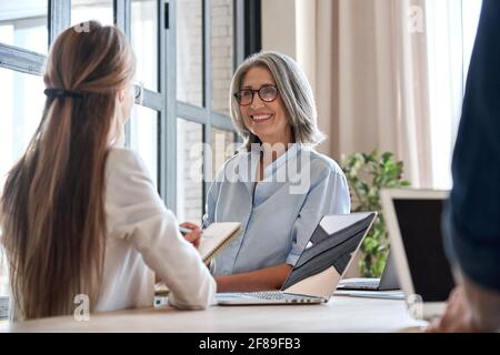 Senior business woman working on laptop with assistant writing notes. Stock Photo