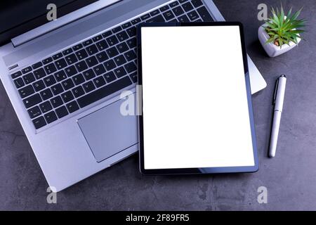 Tablet Pc with white blank screen over silver laptop with pencil, flower on desktop. Stock Photo