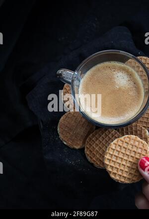 Closeup view of a foamy creamy coffee cup with some chocolate cookies around and a woman hand holding one of them. Stock Photo