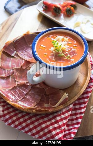 Bowl of delicious cold tomato soup, spanish salmorejo, in a wooden tray full of ham dry-cured ham slices. Stock Photo