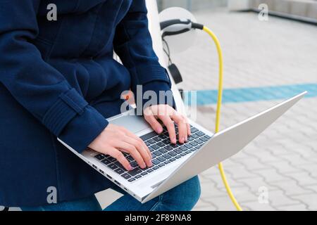 Woman sits on the trunk of a car and uses a notebook while the EV is charging Stock Photo