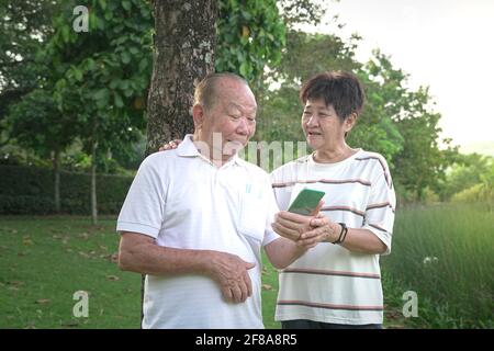 Happy senior Asian couple smiling, talking and looking at phone in the park. Stock Photo