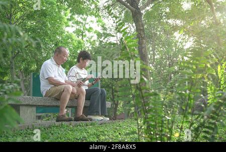 Senior couple sitting on bench, reading their phones in the park. Digital lifestyle of retirees or elderly concept. Stock Photo