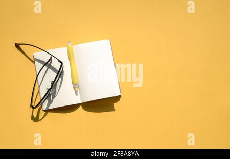 Open notepad, pen and reading glasses on top of yellow background table. Top view, copy space. Stock Photo