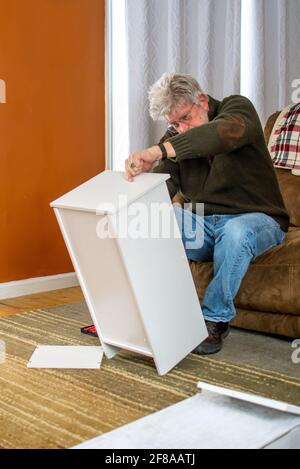 Older male puts a small cabinet together in his living room Stock Photo