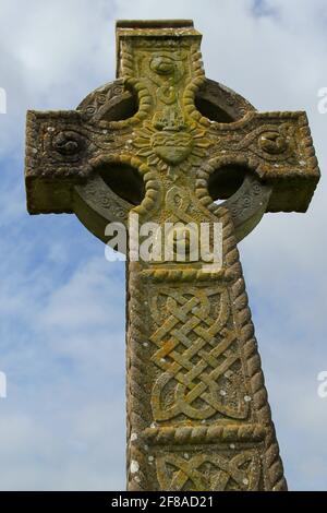 Green Moss Covered Stone Celtic Cross with Heart Carving Against Cloudy Blue Sky in Ireland Stock Photo