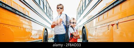 Caucasian brothers students near yellow school bus. Cool kids in sunglasses going back to school in September. Education system and learning. Support Stock Photo