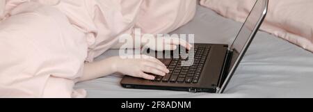 Bored funny child lying in bed under blanket and learning in a virtual online school class. Hands typing on keyboard. Kid working on laptop Internet. Stock Photo