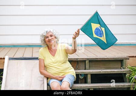 Proud citizen celebrating Independence Day of Brazil. Happy old woman with Brazilian flag outdoor. Smiling Brazilian elderly lady sitting on home back Stock Photo