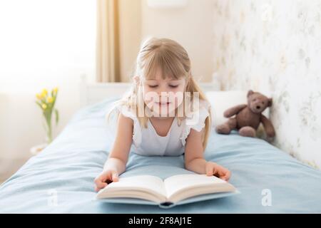 A little girl leys on the bed in the stylish bedroom and reads a blue book, doing homework. Education, home schooling concept. Yellow tulips in the va Stock Photo