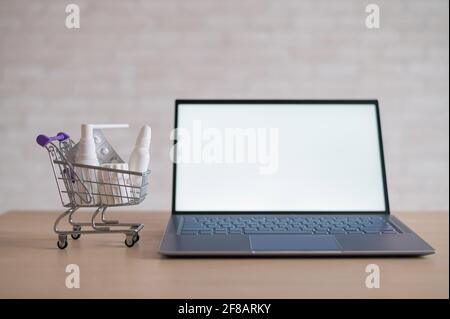Laptop with blank white screen and miniature shopping cart full of medicines. Online pharmacy concept. Website for buying tablets. Stock Photo