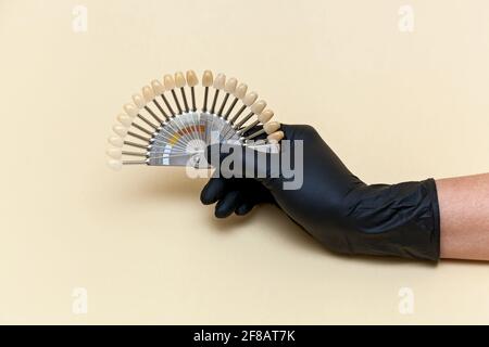 Female hand in a black medical glove holds a coloring scale of artificial teeth for visual measurement of color on a beige background. Concept of dent Stock Photo