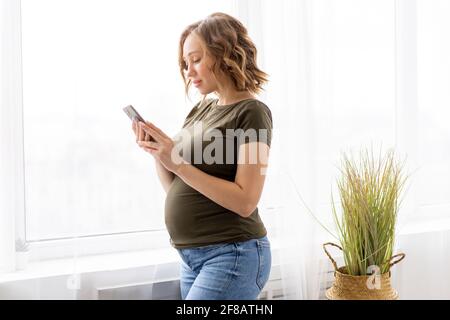 Pregnant woman use smartphone standing near big window Pregnancy Caucasian female with cell phone Modern technology concept Stock Photo