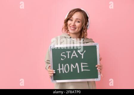 Stay home concept Positive message Woman with headphones dressed oversize hoodie holds chalkboard with the words stay home. Coronavirus protection New Stock Photo