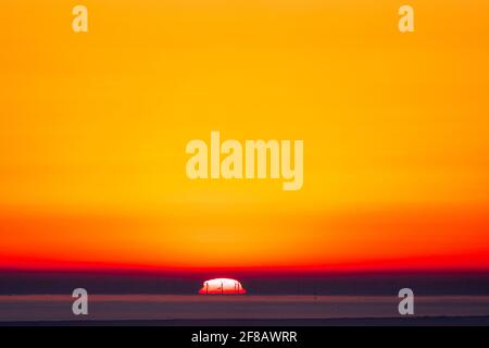 Sunrise over the sea with silhouetted wind turbines in front of the sun. The sun rising over the Thanet Off shore windfarm off the Kent coast in England. Layer of haze on horizon with the turbines showing against the white ball of the sun only. Red and yellow clear sky above. Stock Photo