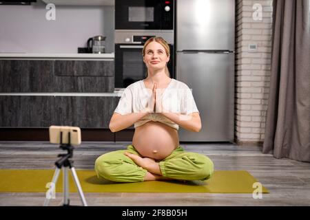 Healthy Pregnant Female taking photo or recording video online, lead translation for subscribers, doing yoga exercises at home, in kitchen. healthy li Stock Photo