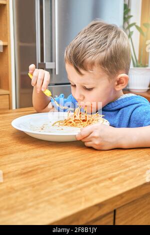 Lovely boy kid with large eyes eats sloppy delicious pasta with yellow baby fork sitting in modern kitchen against metal fridge Stock Photo