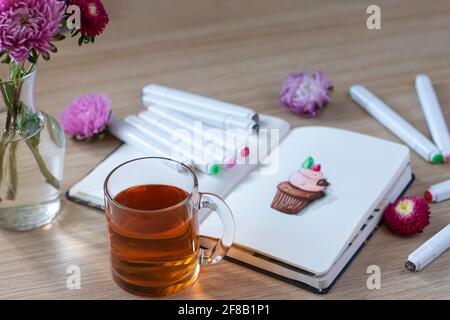 All placed on a wooden table with a transparent cup of tea, notebook, markers, and flowers. sketching cupcake in a notebook. Top view Stock Photo