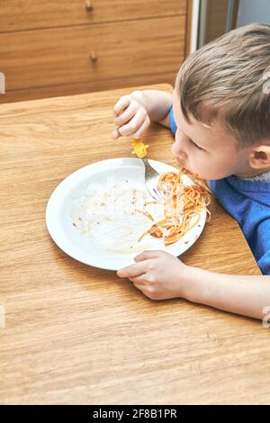 Lovely boy kid with large eyes eats sloppy delicious pasta with yellow baby fork sitting in modern kitchen against Stock Photo