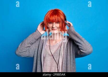 teenage girl in headphones listens to music on blue background. girl with bright red hair in anime style Stock Photo
