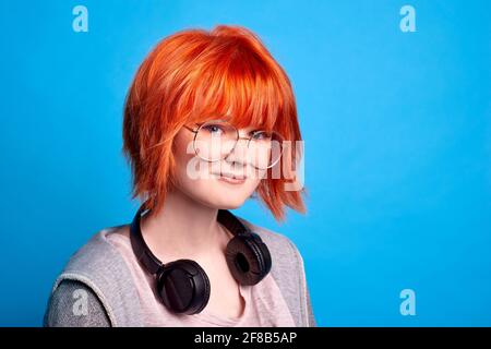 hipster girl with wireless headphones on blue background. smiling teenager with red hair Stock Photo